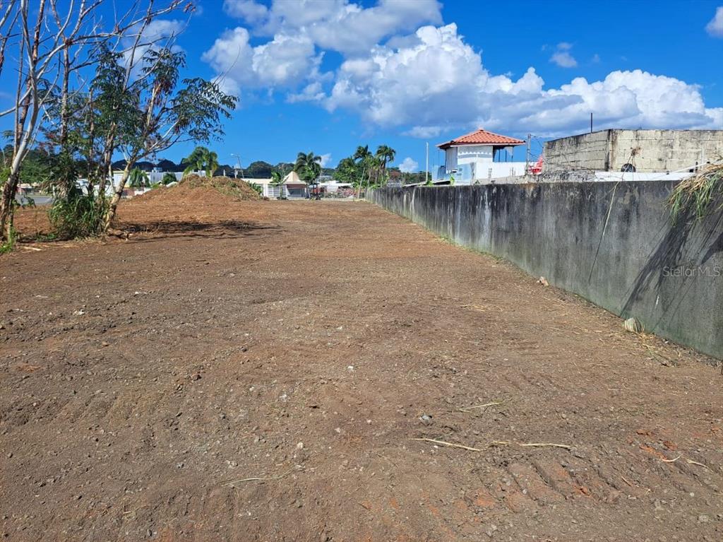 A cleared, vacant lot of dirt features a tall concrete wall along the right side — CARRETERA #2 CANDELARIA WARD INTERIOR, TOA BAJA, PR