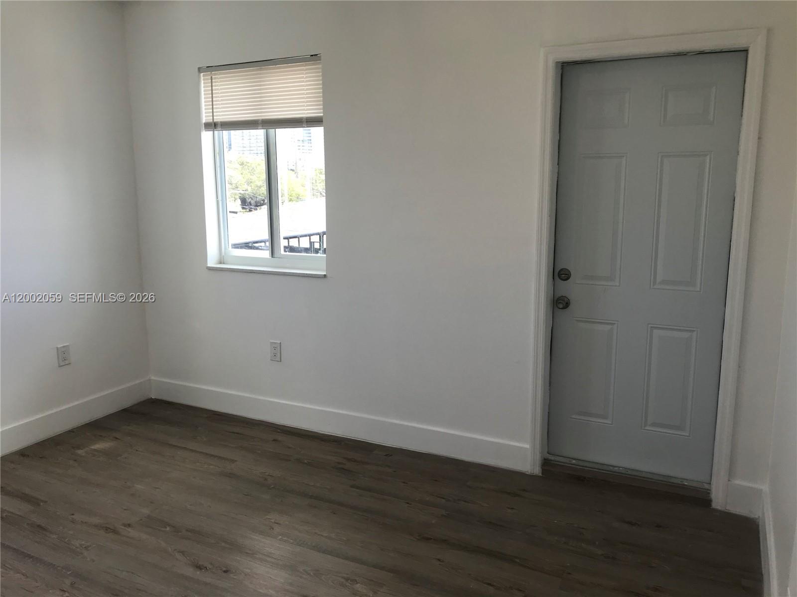 A room with white walls, wood-look flooring, a window with blinds, and a gray door — 1815 NW 1st CT # 8, Miami FL 33136, MIAMI, FL
