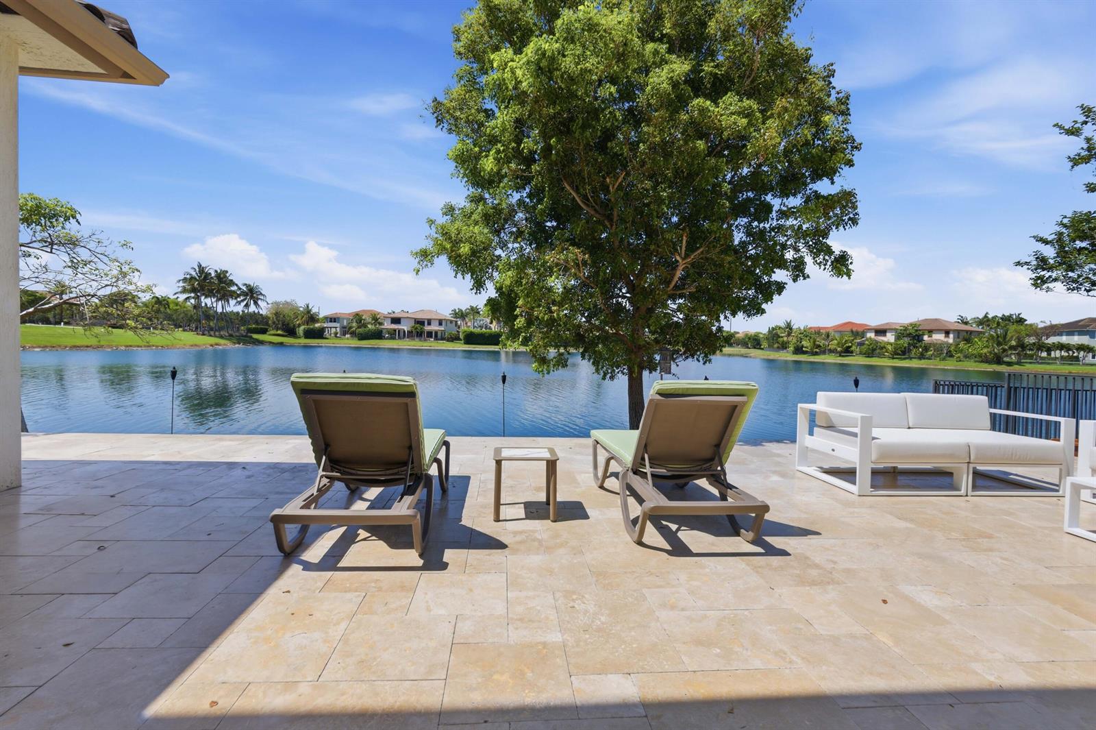 A stone patio features two lounge chairs and a sofa overlooking a lake and trees — 10271 Peninsula Place, Parkland FL 33076, PARKLAND, FL