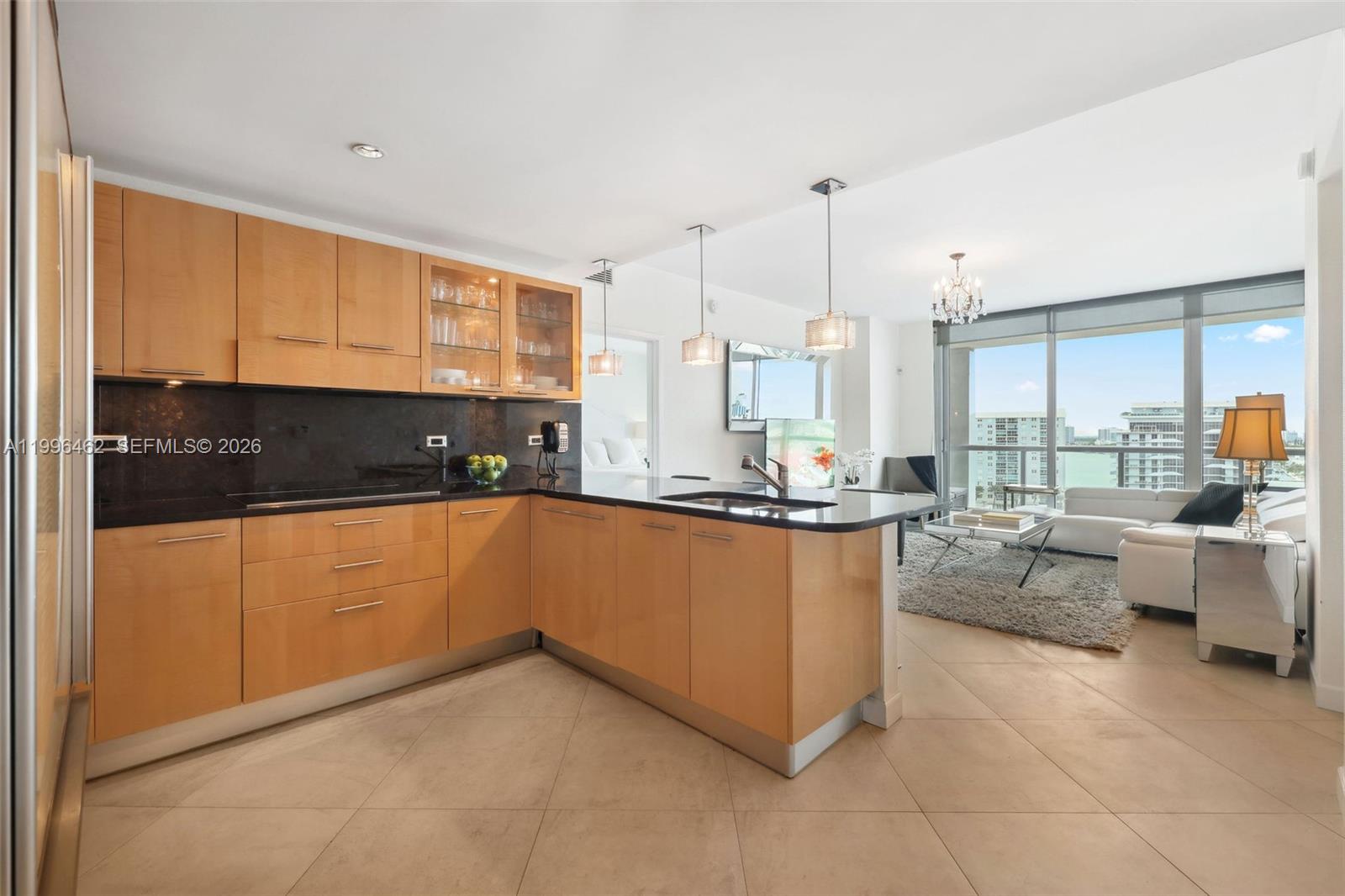 A modern kitchen with light wood cabinets and black countertops opens into a living area — 6799 Collins Ave # 1106, Miami Beach FL 33141, MIAMI BEACH, FL