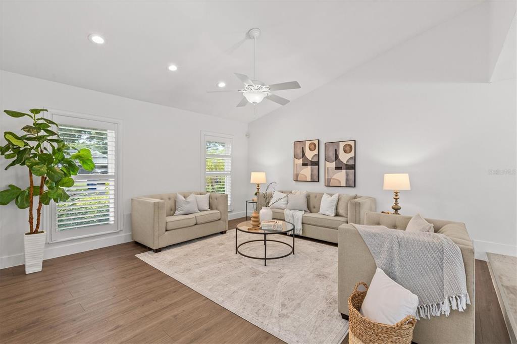 A bright living room features a vaulted ceiling, light-colored sofas, a coffee table, and wood-look flooring — 13915 WHISPERWOOD DRIVE, CLEARWATER, FL
