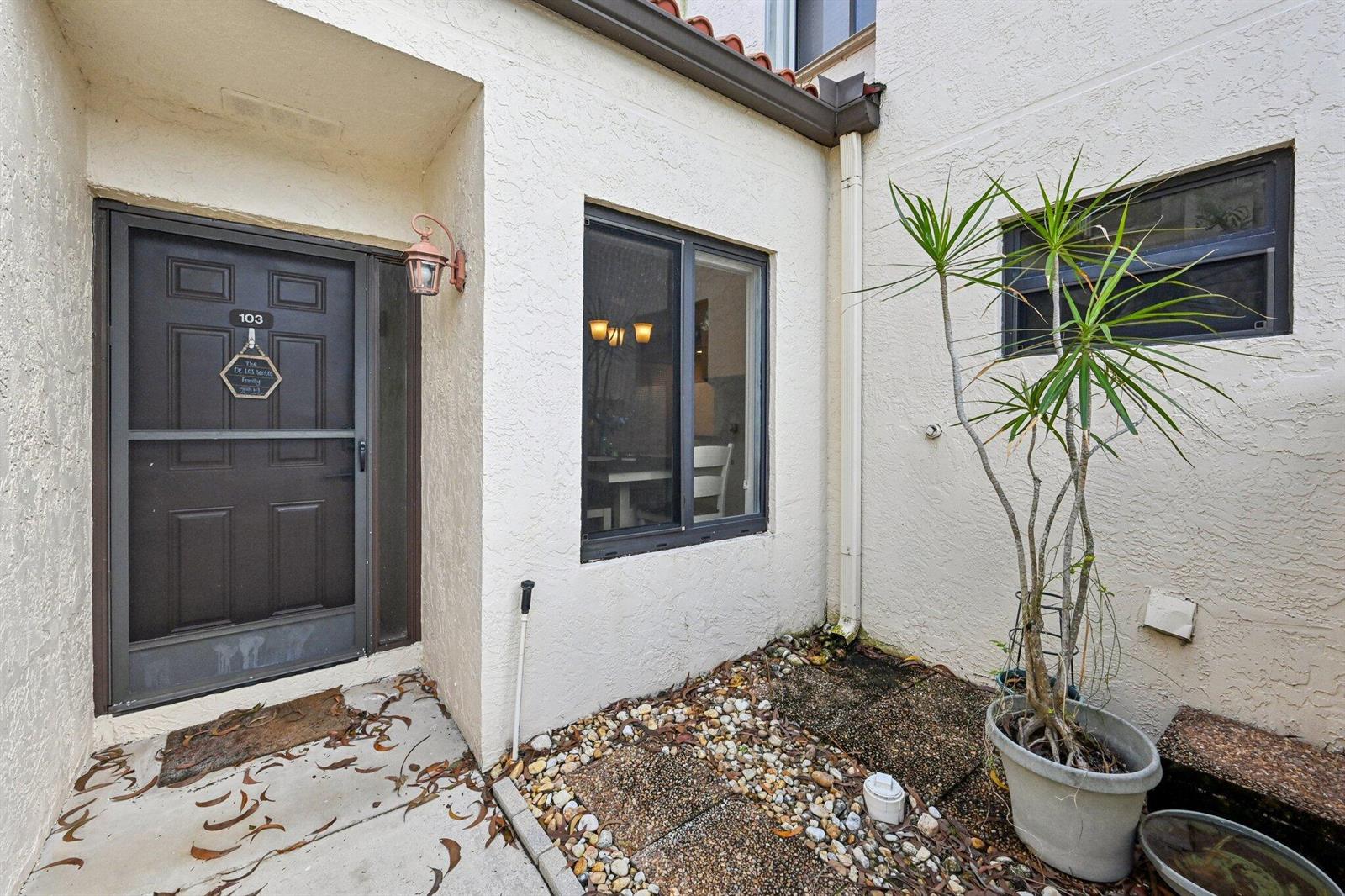 An exterior view of a residential entryway with a dark door, window, and potted plant — 22 Via De Casas # 103, Boynton Beach FL 33426, BOYNTON BEACH, FL