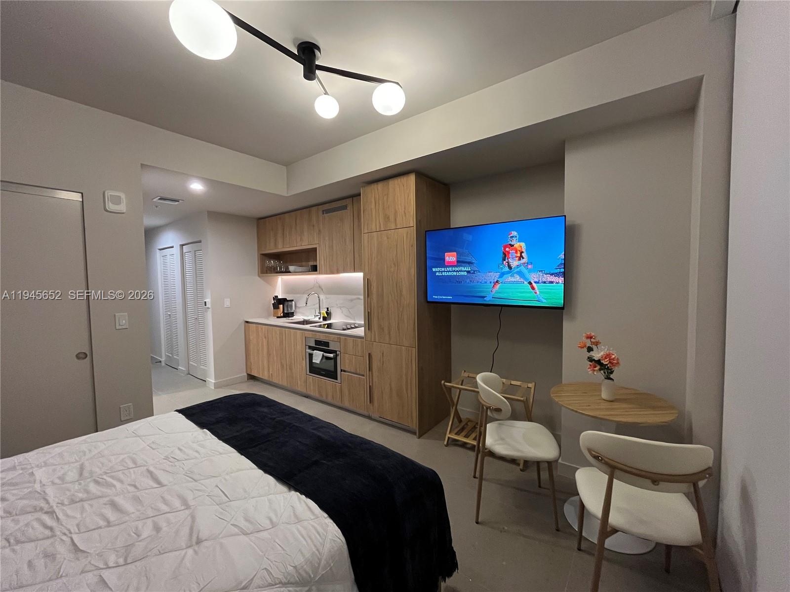 A studio apartment features a kitchenette with wood cabinetry, a bed, and a wall-mounted television — 11 NE 6TH # 1006, Miami FL 33132, MIAMI, FL