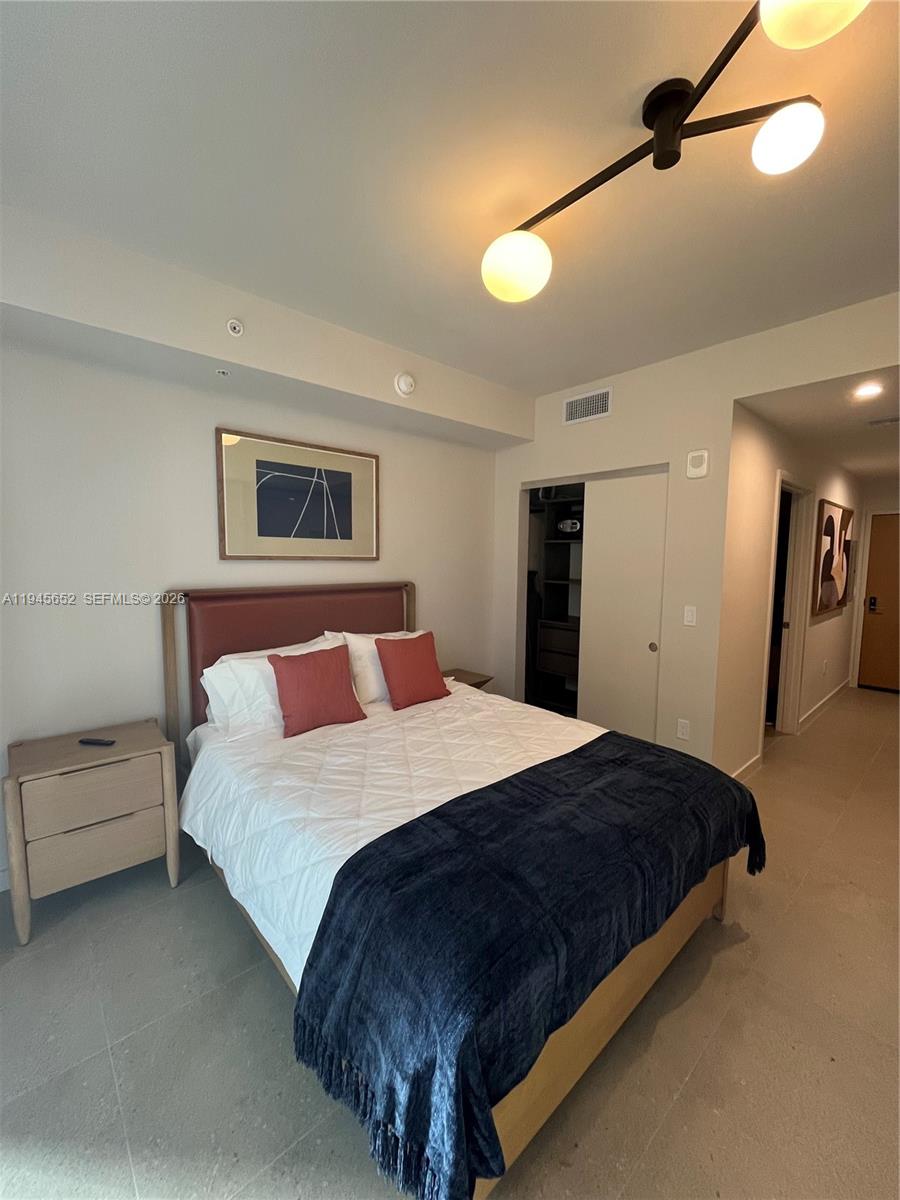 A bedroom features a bed with a dark blue throw, light wood nightstands, and a modern light — 11 NE 6TH # 1006, Miami FL 33132, MIAMI, FL