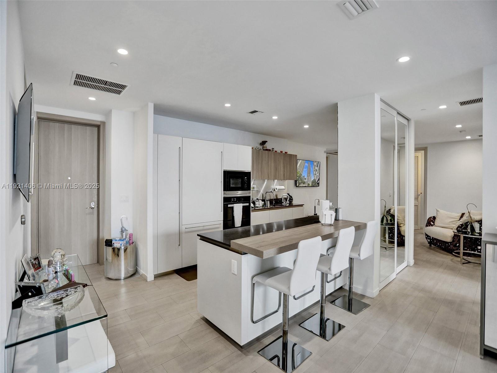 A modern kitchen features white cabinetry, a wood-topped island with bar stools, and light flooring — 1000 Brickell Plz, Miami FL 33131, MIAMI, FL