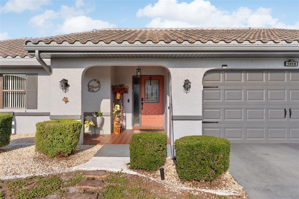 The exterior of a single-story home features a tiled roof, a coral-colored door, and landscaping — 1816 GRANVILLE LANE, SUN CITY CENTER, FL