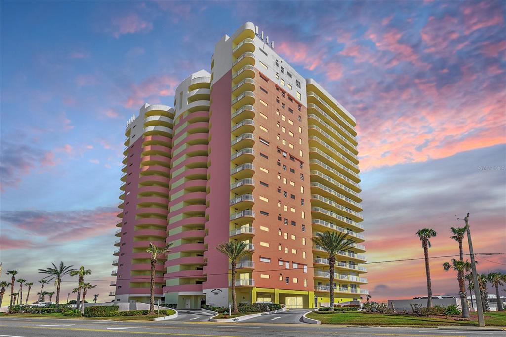 A tall, multi-colored residential high-rise building stands against a sunset sky — 1900 N ATLANTIC AVENUE UNIT 1603, DAYTONA BEACH, FL