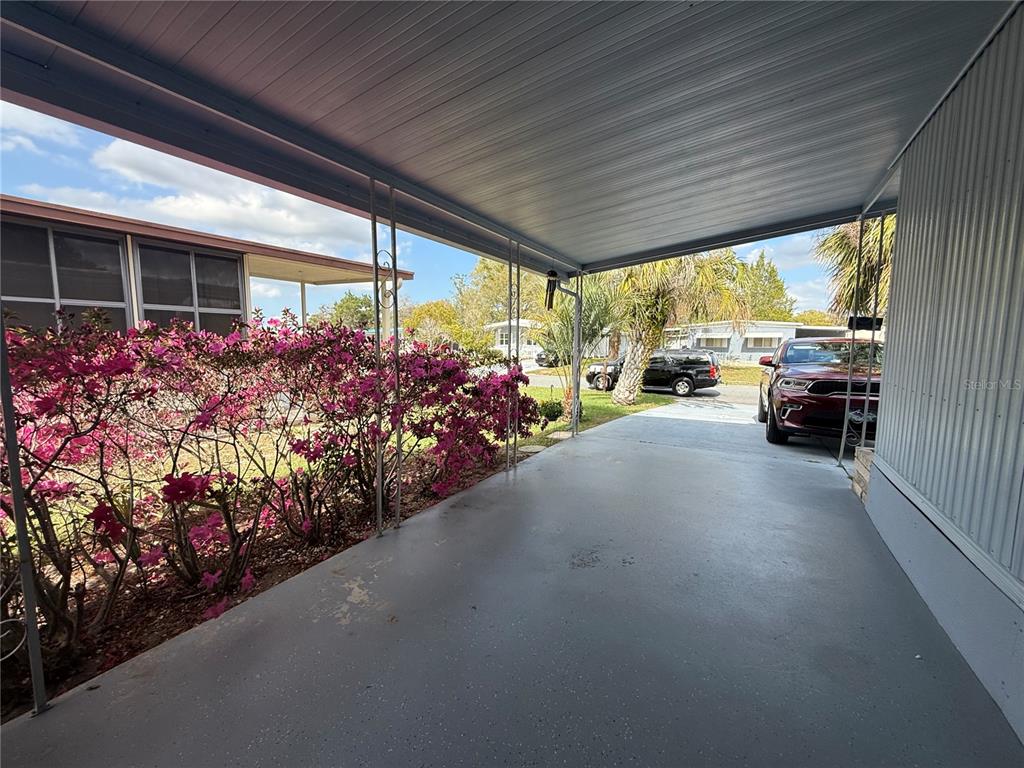 A covered carport features a concrete floor, metal support poles, and adjacent blooming azalea bushes — 7441 FAIRLANE AVENUE, BROOKSVILLE, FL