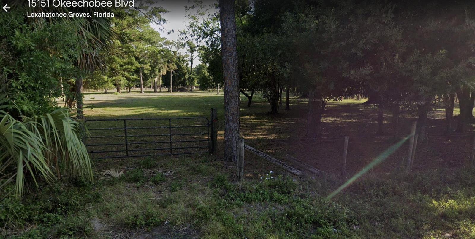 A metal gate opens into a grassy, tree-filled lot with a wire fence — 15151 Okeechobee Boulevard, FL 33470, FL