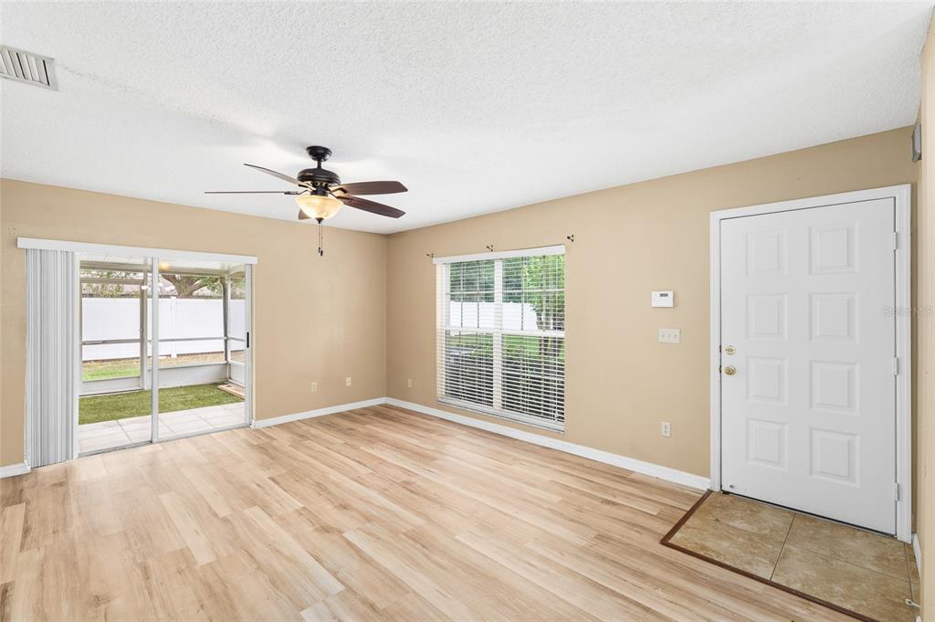 A living room with light wood-look flooring, a ceiling fan, sliding doors, and a front door — 539 KENSINGTON LAKE CIRCLE, BRANDON, FL