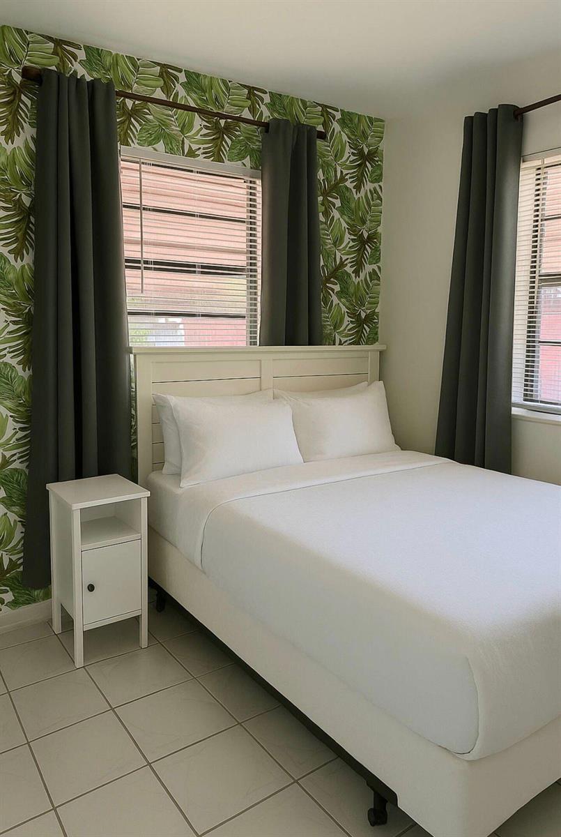 A bedroom features a white bed, a nightstand, and walls with tropical leaf wallpaper — 272 NE 44th Street, Deerfield Beach FL 33064, DEERFIELD BEACH, FL