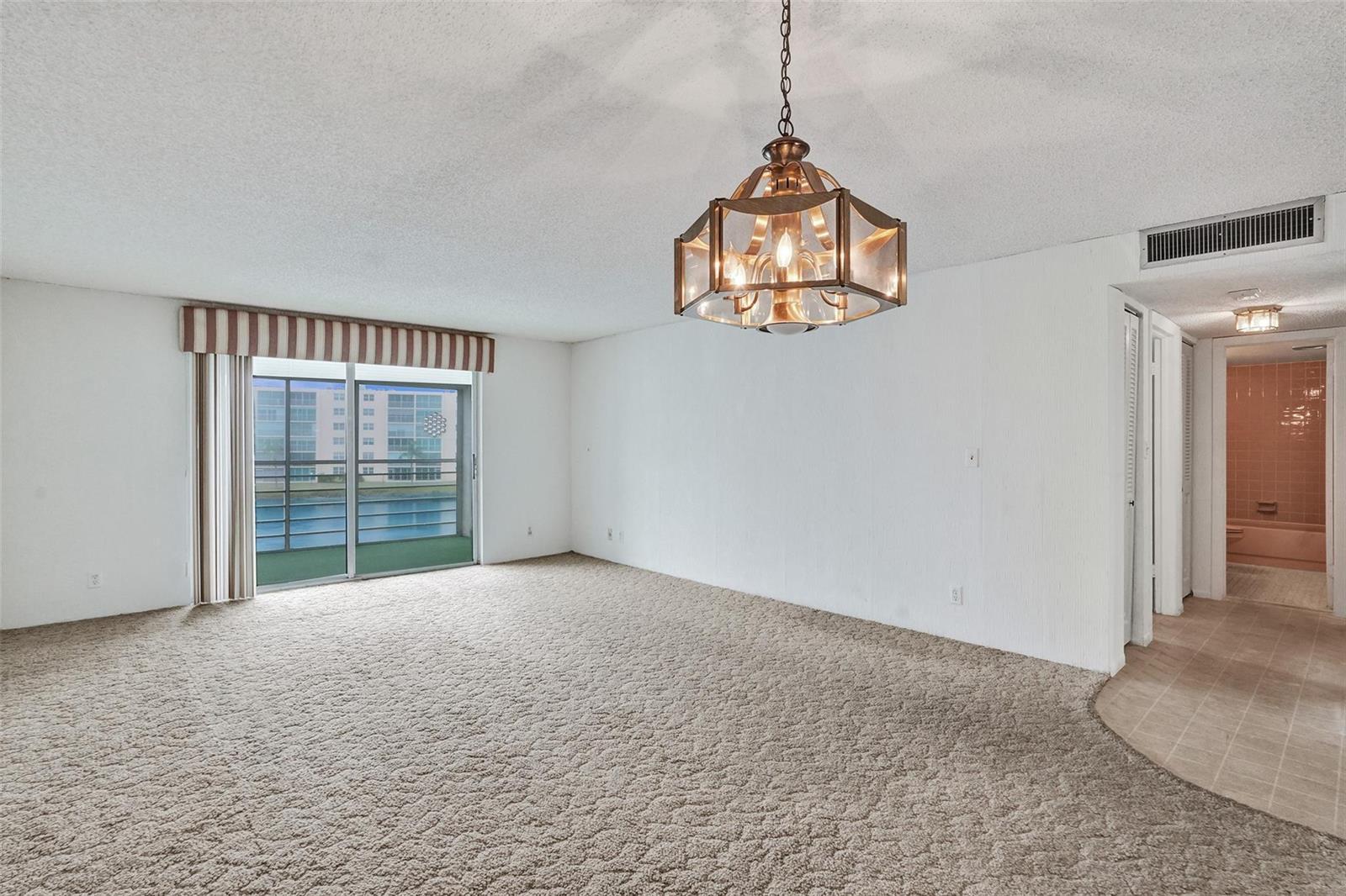 An empty living room features beige wall-to-wall carpeting, a decorative chandelier, and a sliding glass door — 131 SE 3rd Avenue # 304, Dania Beach FL 33004, DANIA BEACH, FL