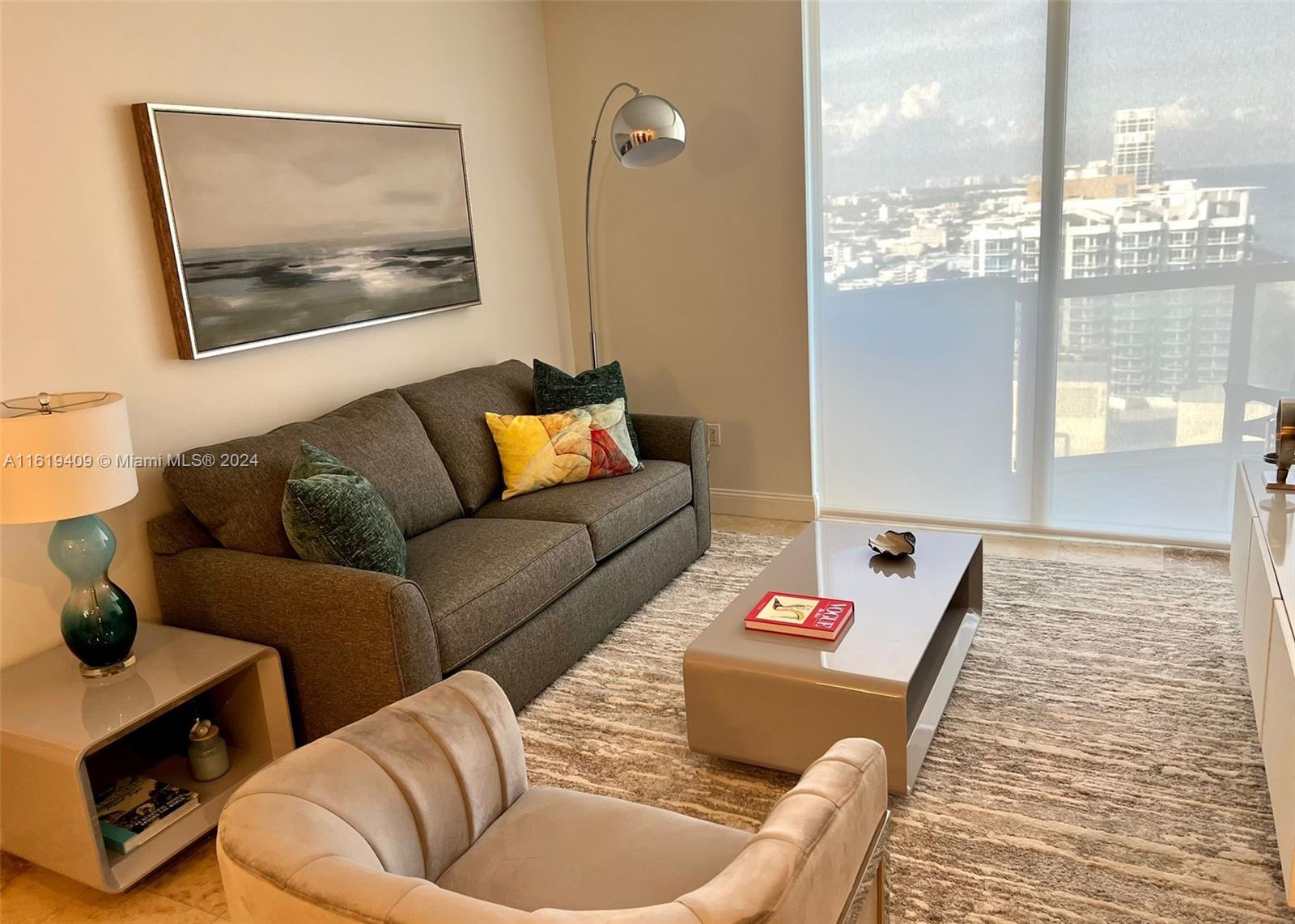 A living room features a gray sofa, a beige armchair, and a view of city buildings — 6365 Collins Ave # 2610, Miami Beach FL 33141, MIAMI BEACH, FL