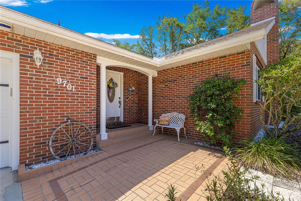 A brick house exterior features a covered entryway with a white bench and paved patio — 9701 SW 200TH COURT, DUNNELLON, FL