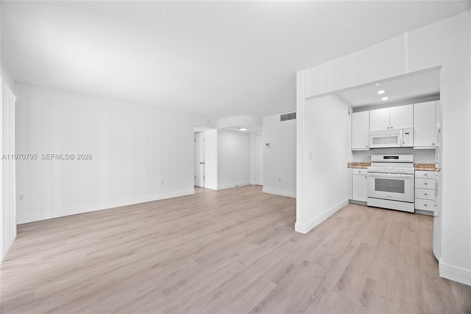 An open living area with light wood-look flooring leads into a white kitchen — 4990 E Sabal Palm Blvd # 202, Tamarac FL 33319, TAMARAC, FL