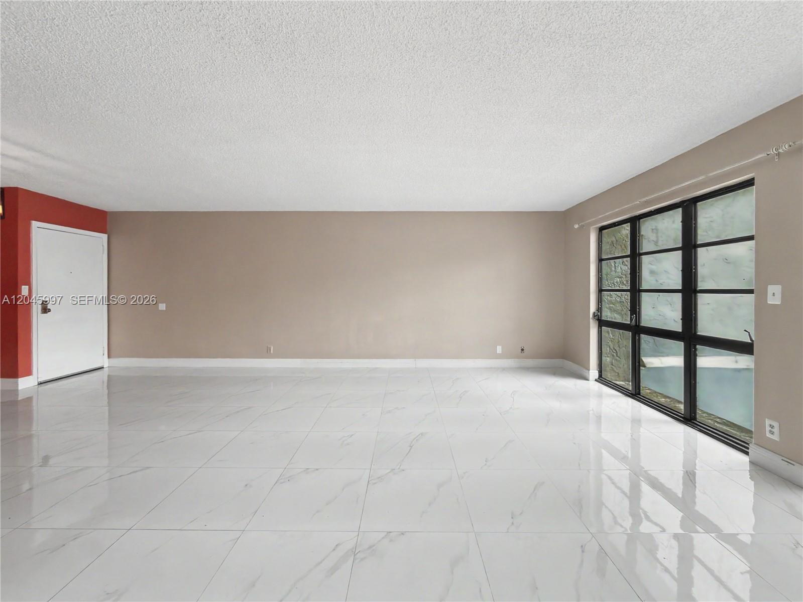 An empty room features white marble-look tile flooring, beige walls, a red accent wall, and windows — 14203 SW 66th St # 103, Miami FL 33183, MIAMI, FL