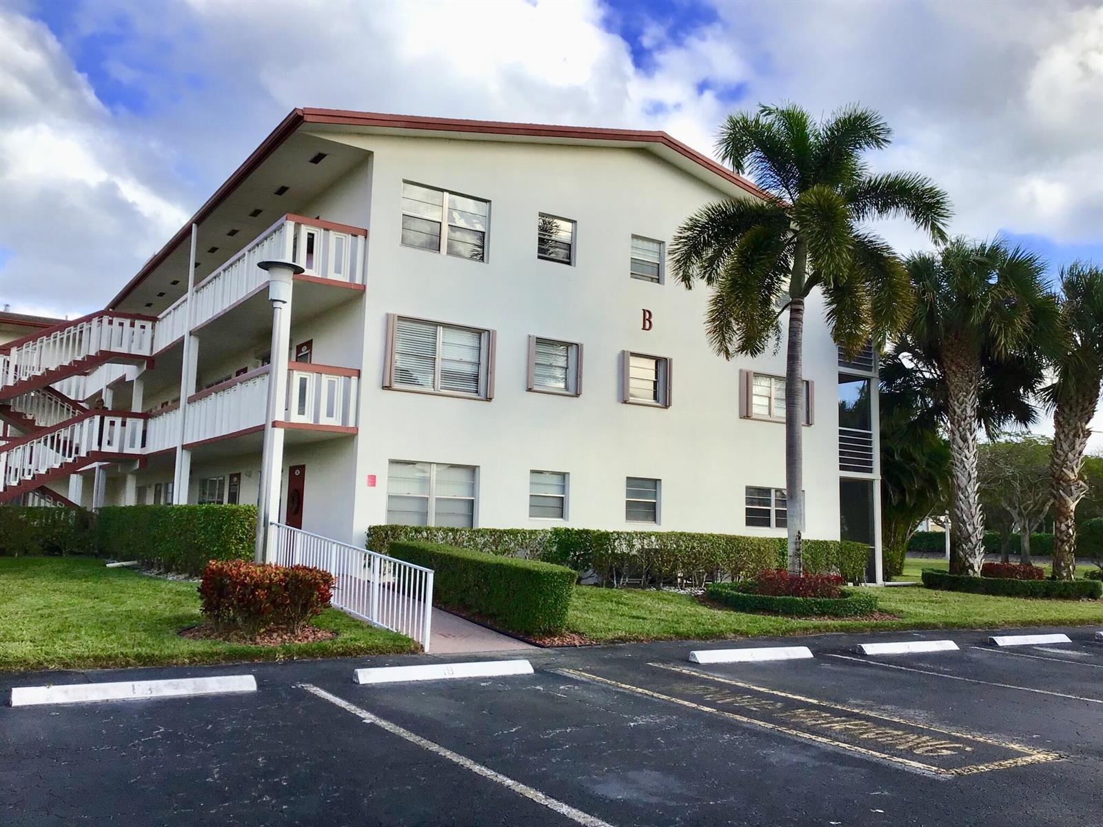 A three-story apartment building with white stucco walls, balconies, and palm trees in the foreground — 84 Brighton B, Boca Raton FL 33434, BOCA RATON, FL