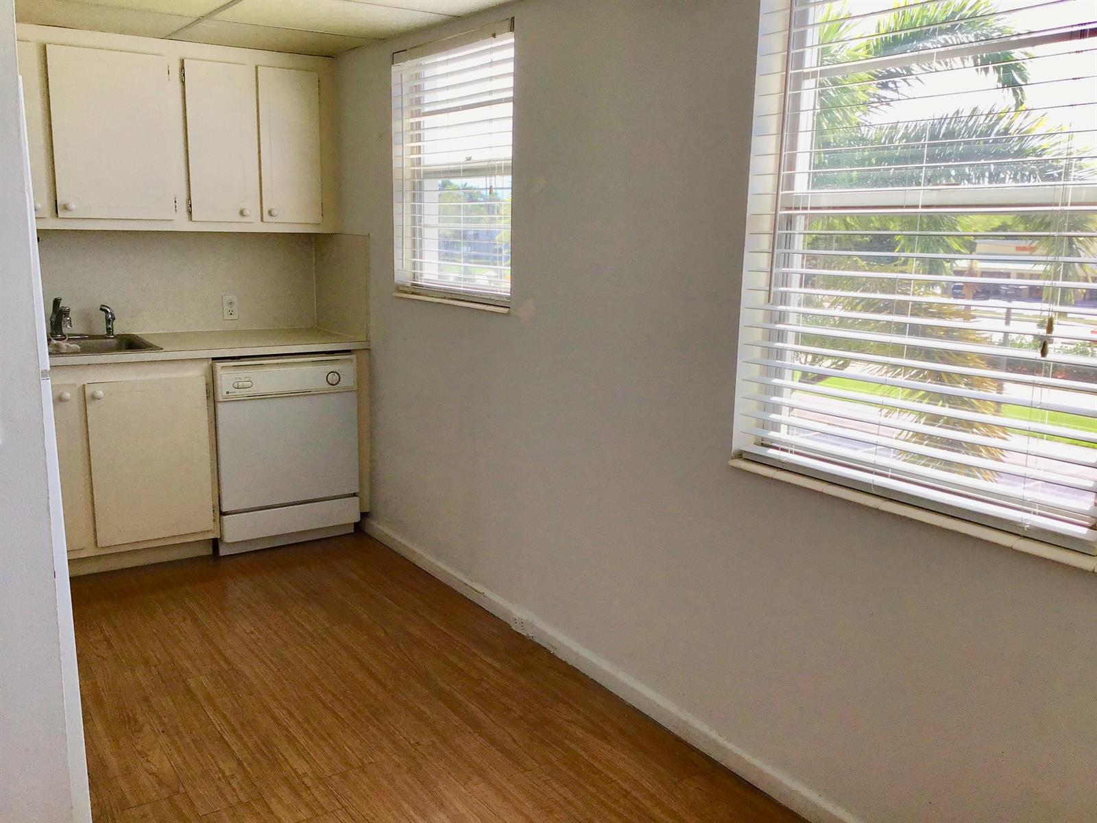A small kitchen features white cabinets, a dishwasher, a sink, and wood-look flooring — 84 Brighton B, Boca Raton FL 33434, BOCA RATON, FL