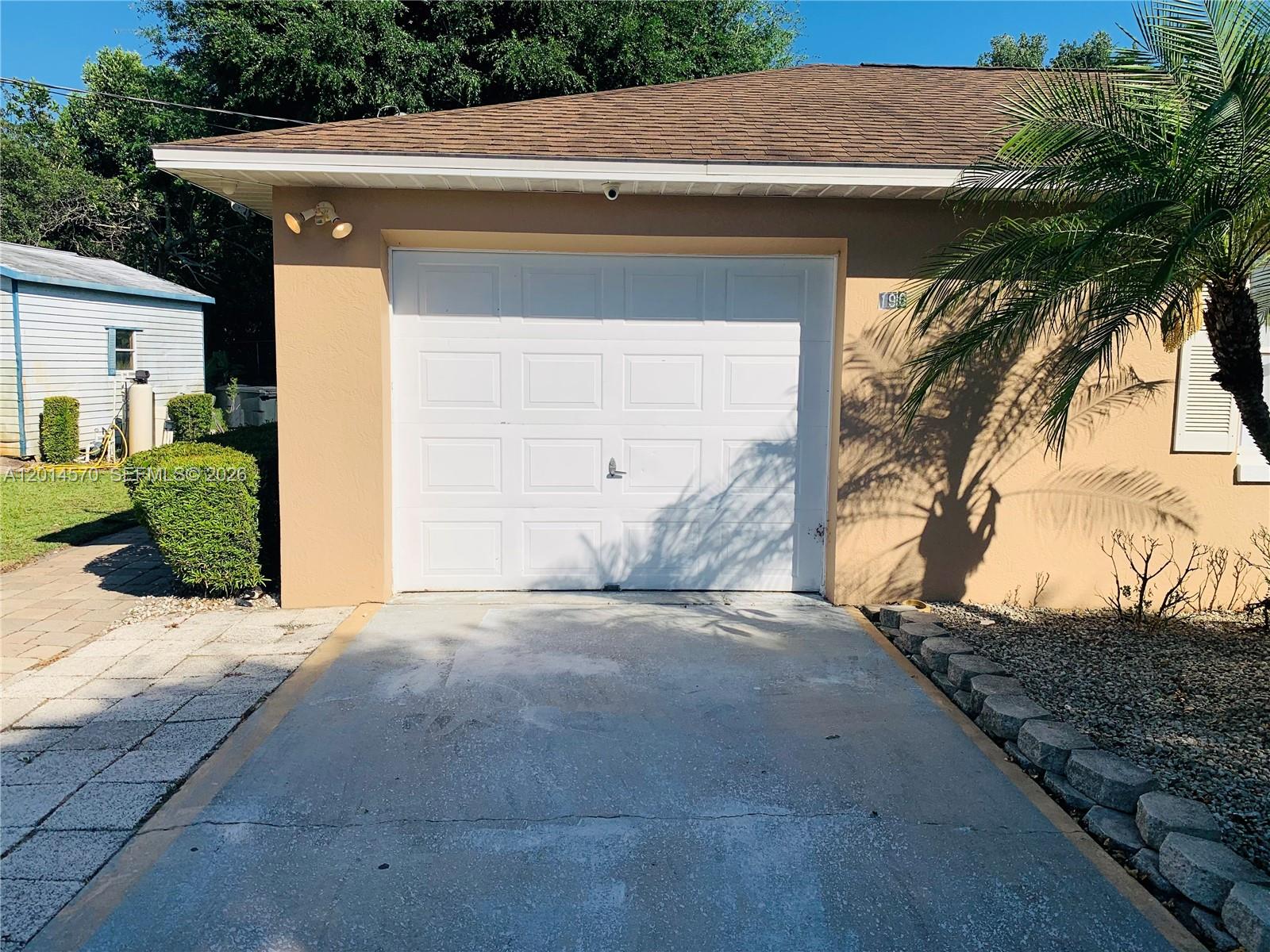 A tan garage with a white door and a concrete driveway sits under a palm tree — 196 JAMISON AVE, Lake Placid FL 33852, LAKE PLACID, FL