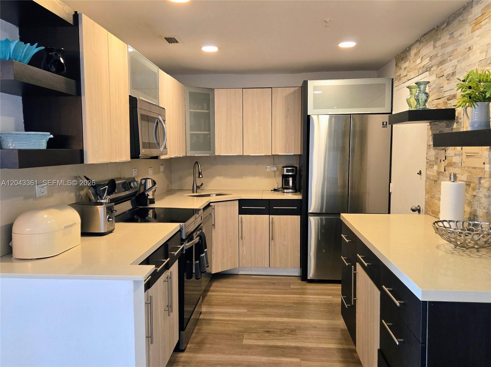 A modern kitchen features light wood cabinets, dark accents, stainless steel appliances, and stone wall tiling — 5601 Collins Ave # 520, Miami Beach FL 33140, MIAMI BEACH, FL