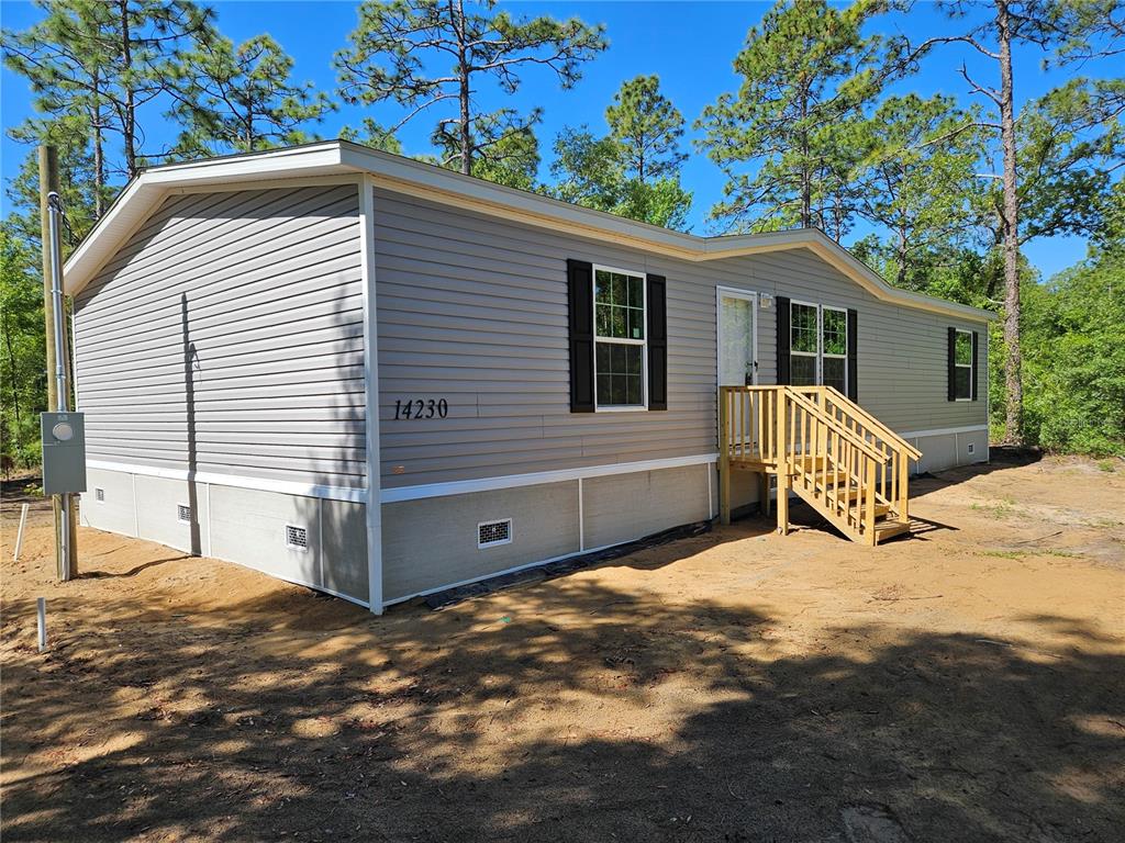 A single-story manufactured home with grey vinyl siding and a wooden staircase sits on dirt — 14230 SE 20TH STREET, MORRISTON, FL