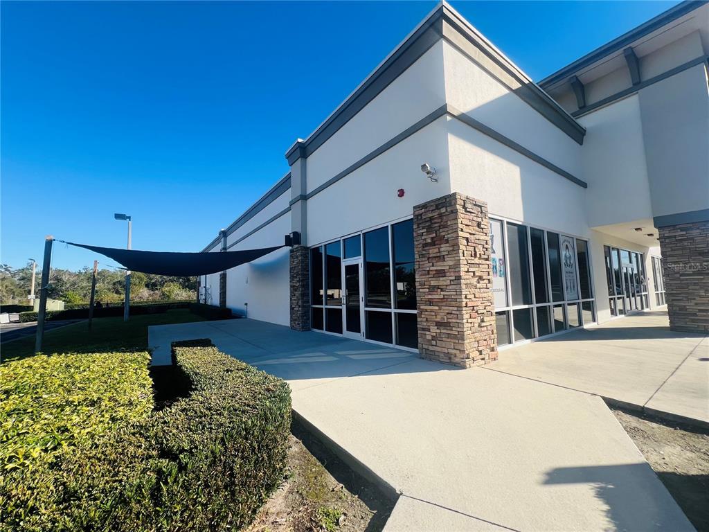 A commercial building exterior features a stone-accented corner, glass storefront windows, and a shade sail — 2507 LAKEWOOD RANCH BOULEVARD, BRADENTON, FL