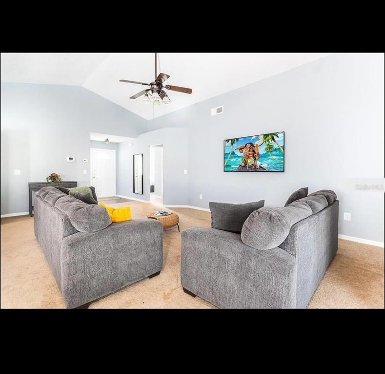 A living room features vaulted ceilings, carpeted flooring, two gray sofas, and a wall-mounted television — 2609 MONTEGO BAY BOULEVARD, KISSIMMEE, FL