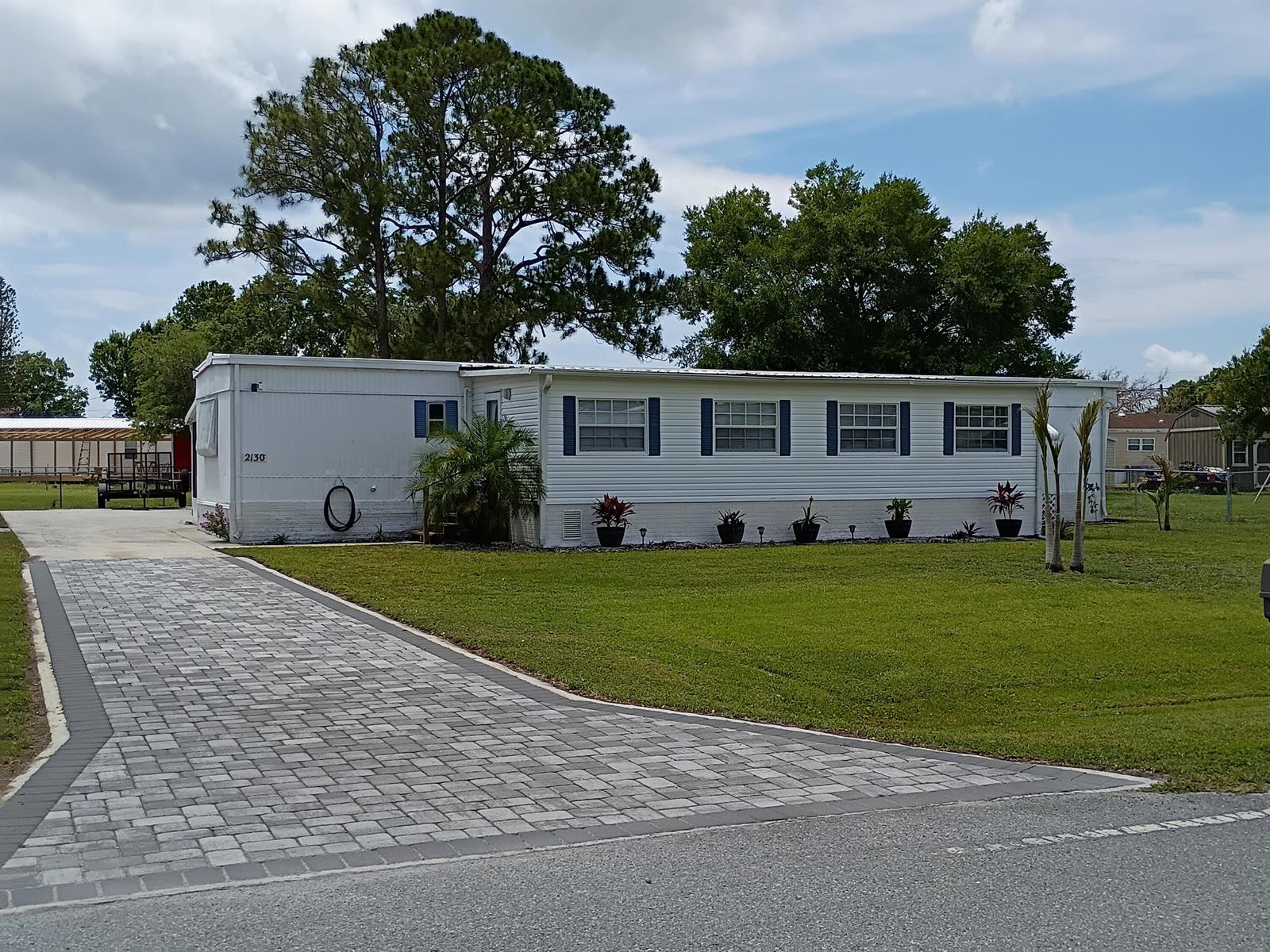 A white mobile home with blue shutters features a paved driveway and a grassy lawn — 2130 SE 24th Boulevard, Okeechobee FL 34974, OKEECHOBEE, FL