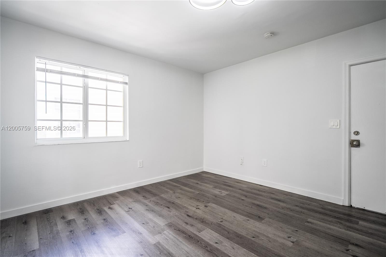An empty room features white walls, dark wood-look flooring, a window, and a door — 920 Pennsylvania Ave # 206, Miami Beach FL 33139, MIAMI BEACH, FL
