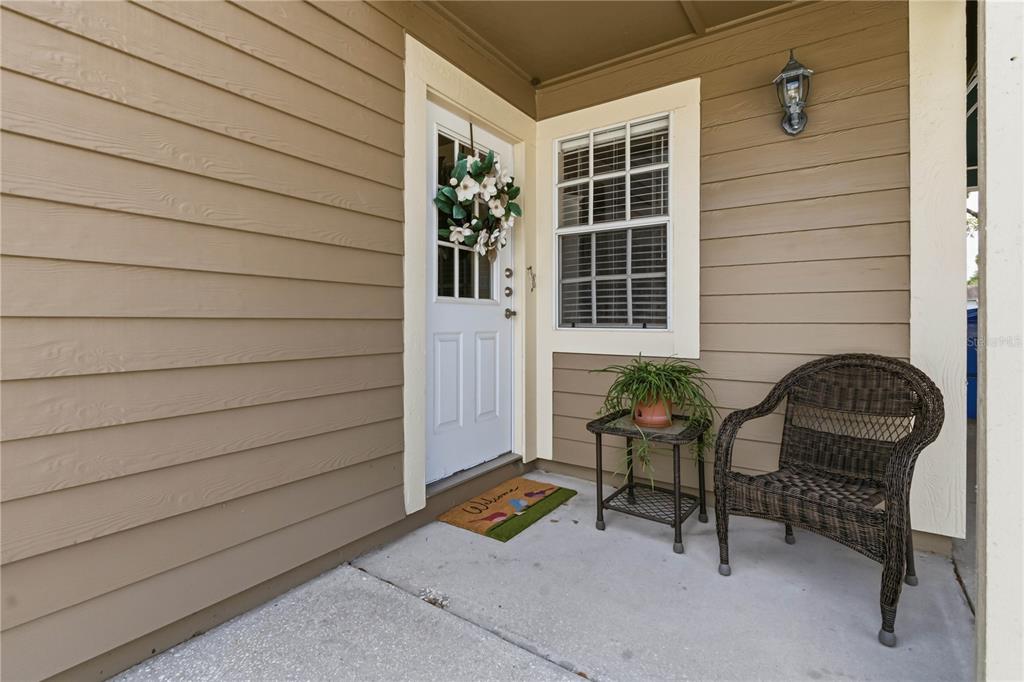 A front porch features tan horizontal siding, a white door with a wreath, and furniture — 9034 LAKE PLACE LANE UNIT 9034, TAMPA, FL