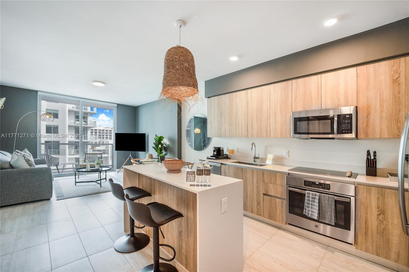 A modern kitchen with light wood cabinets and a breakfast bar opens to a living area — 3900 Biscayne Blvd # 1016, Miami FL 33137, MIAMI, FL