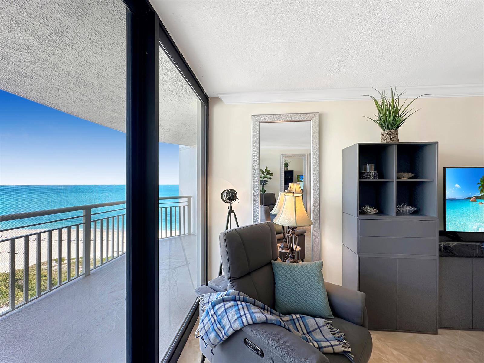 A living area features a gray recliner, a tall shelving unit, and a balcony overlooking ocean — 3000 N Ocean Drive # B, Singer Island FL 33404, SINGER ISLAND, FL