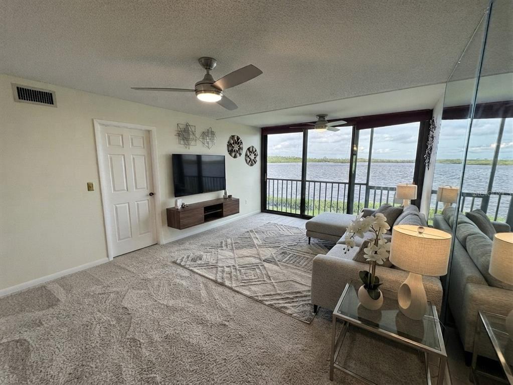 A living room features gray carpeting, a wall-mounted television, and a balcony overlooking the water — 4620 BAY BOULEVARD UNIT 1112, PORT RICHEY, FL
