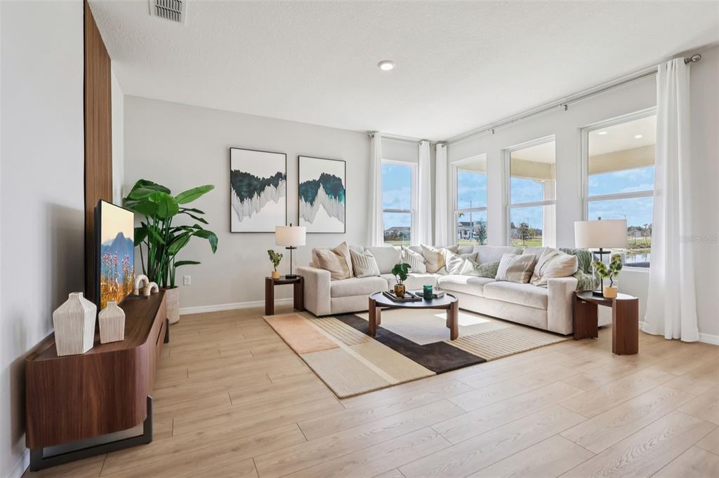 A bright living room features a sectional sofa, wood-look flooring, and large windows with curtains — 5308 CLOUDBERRY AVENUE, LAKELAND, FL