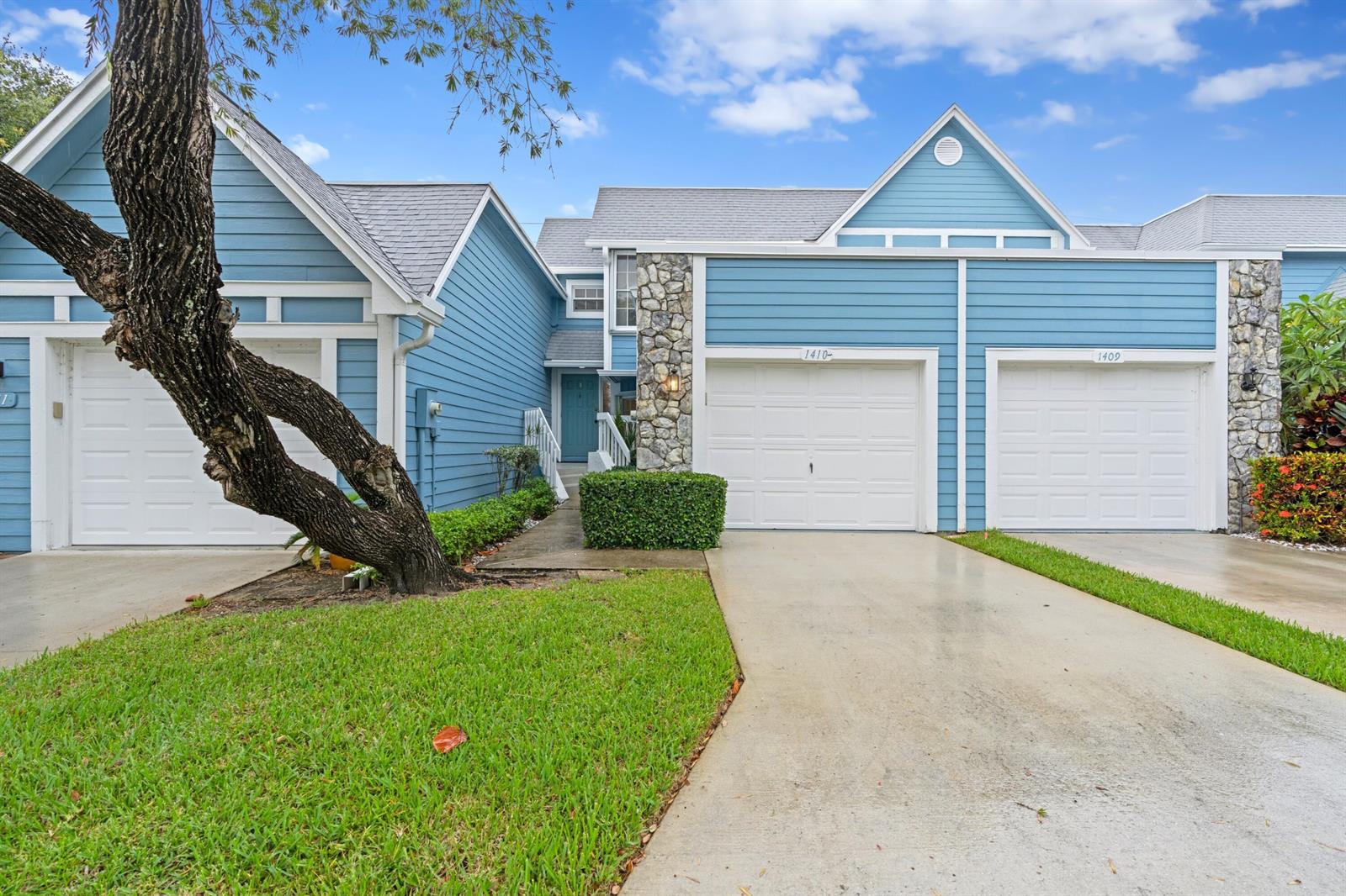 A blue-sided townhouse exterior features two garage doors, a stone facade, and a large tree — 1410 Ocean Dunes Circle, Jupiter FL 33477, JUPITER, FL