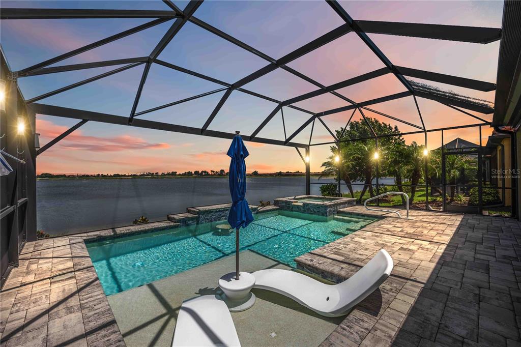 A screened-in patio features a swimming pool, a hot tub, and lounge chairs overlooking a lake — 24100 SPARTINA DRIVE, VENICE, FL