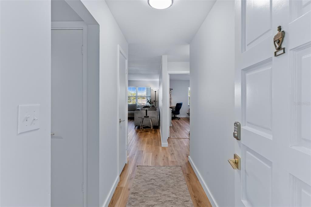 A hallway with light wood flooring leads from a white door into a living area — 701 POINSETTIA ROAD UNIT 208, BELLEAIR, FL