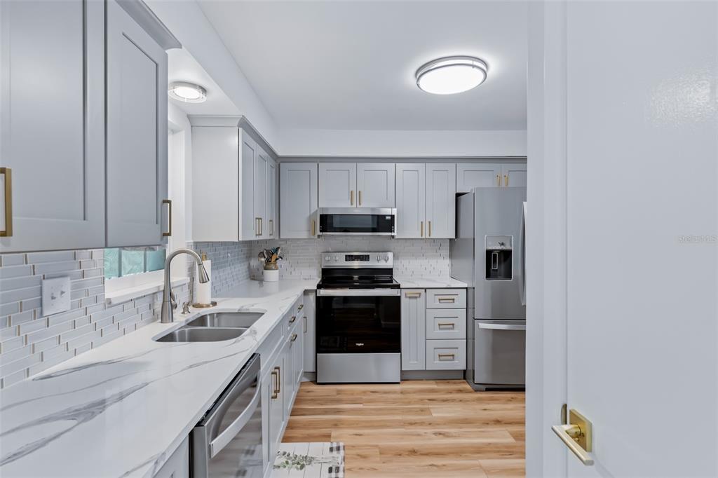 A kitchen features gray cabinets, stainless steel appliances, white marble-patterned countertops, and light wood flooring — 701 POINSETTIA ROAD UNIT 208, BELLEAIR, FL
