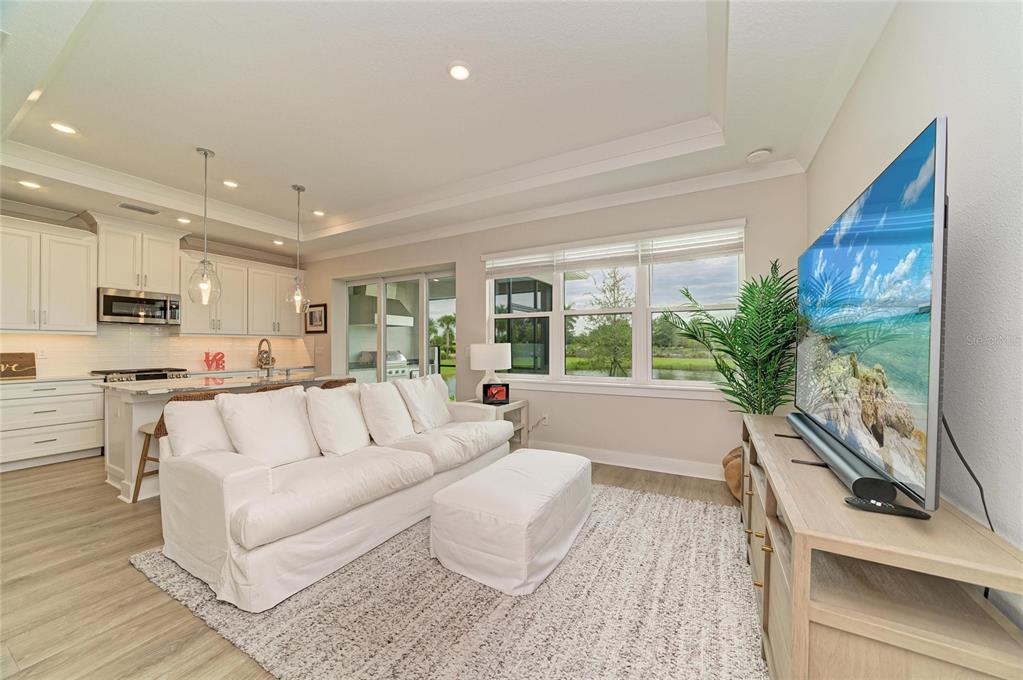 A living room with a white sofa, ottoman, and television, open to a kitchen area — 4064 SANTA CATERINA BOULEVARD UNIT 101, LAKEWOOD RANCH, FL