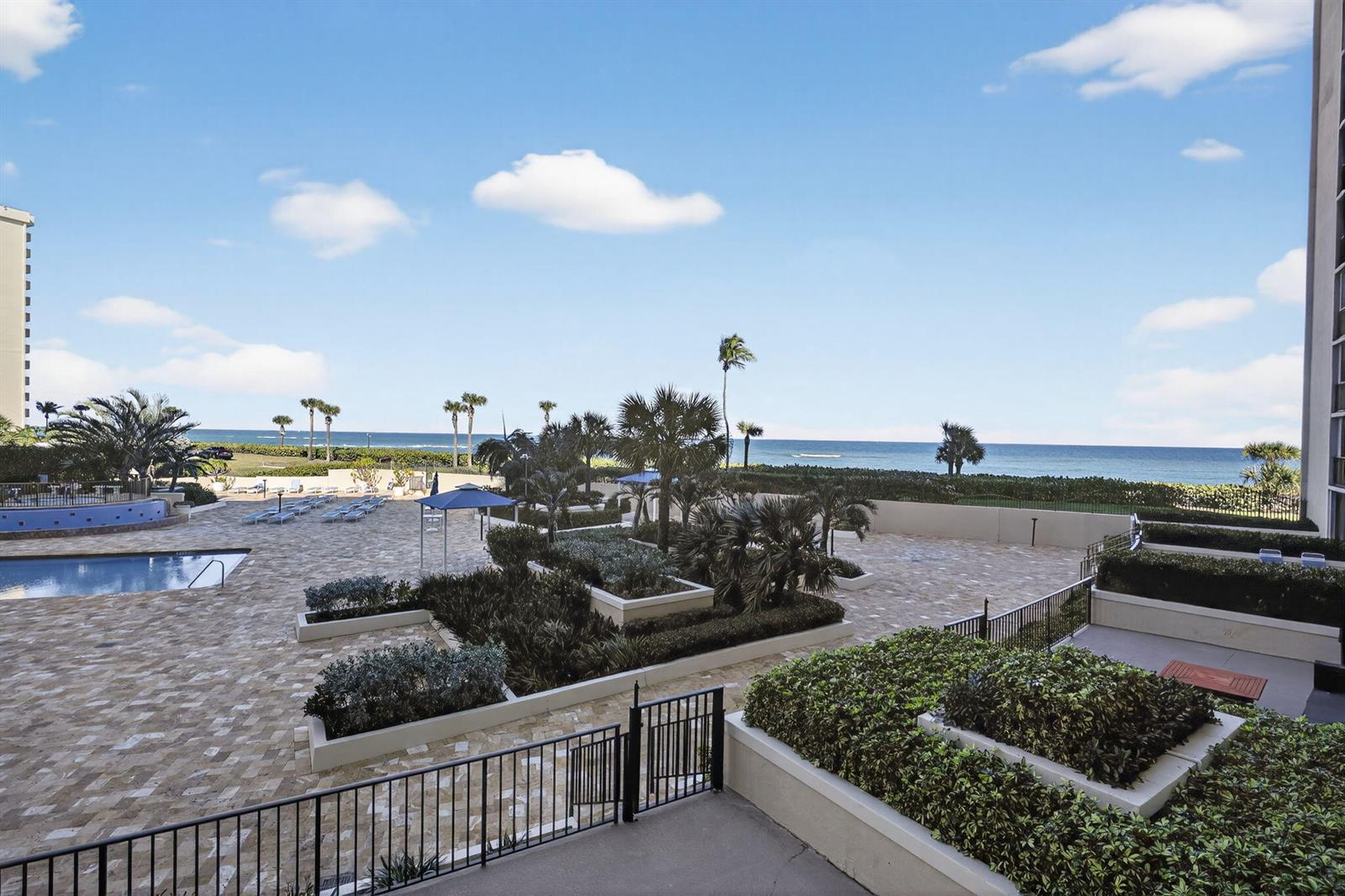 An elevated view of a paved pool deck with landscaping, palm trees, and ocean views — 100 Ocean Trail Way # 204, Jupiter FL 33477, JUPITER, FL