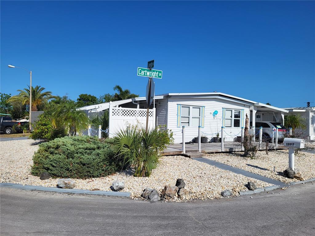A white mobile home with a wooden deck sits on a corner lot with landscaping — 12017 CARTWRIGHT DRIVE, HUDSON, FL