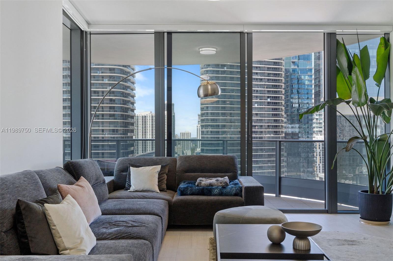 A living room features a grey sectional sofa, a floor lamp, and floor-to-ceiling city views — 1000 Brickell Plz # 2812, Miami FL 33131, MIAMI, FL