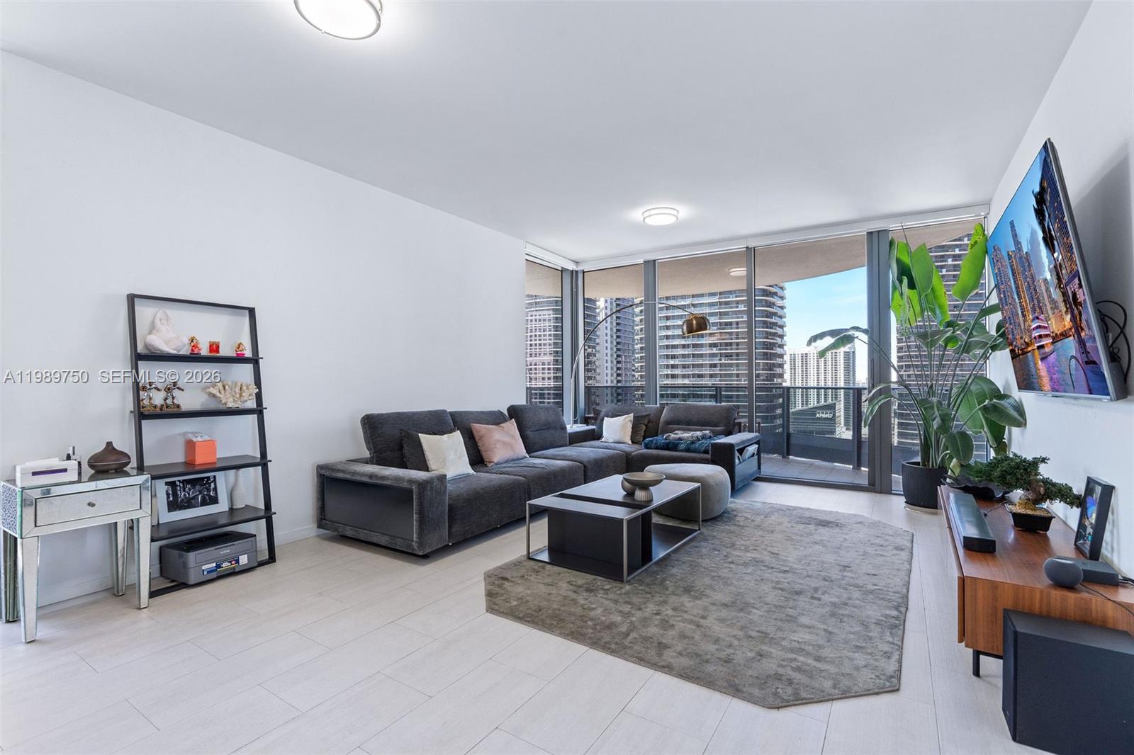 A modern living room features a gray sectional sofa, a television, and floor-to-ceiling windows — 1000 Brickell Plz # 2812, Miami FL 33131, MIAMI, FL