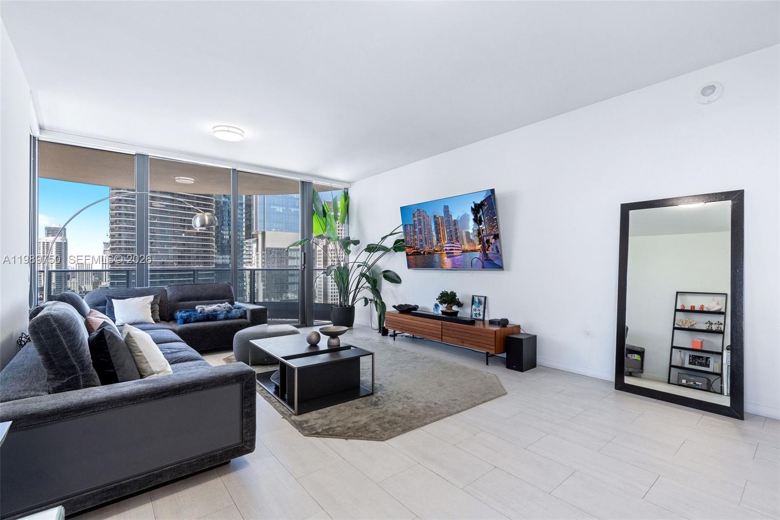 A modern living room features a grey sectional sofa, a television, and a large floor mirror — 1000 Brickell Plz # 2812, Miami FL 33131, MIAMI, FL
