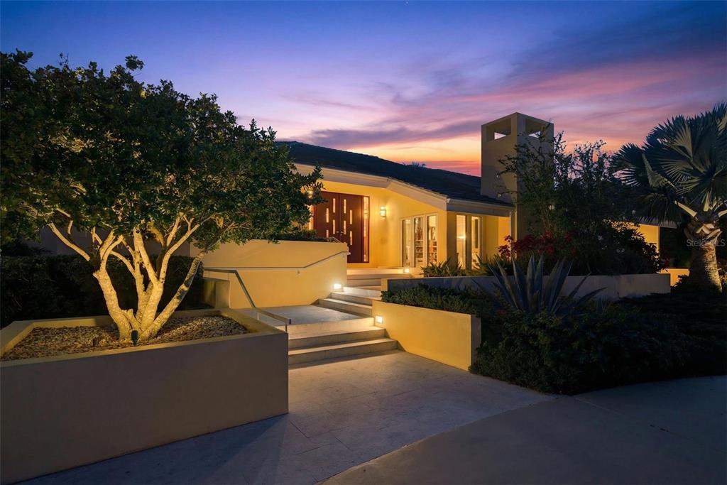 A modern house exterior with a lit entrance, stone steps, and landscaping at dusk — 1571 HARBOR CAY LANE, LONGBOAT KEY, FL