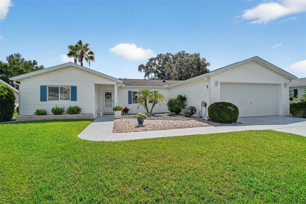 A single-story house with white siding, blue shutters, a two-car garage, and a gravel yard — 17551 SE 96TH COURT, SUMMERFIELD, FL