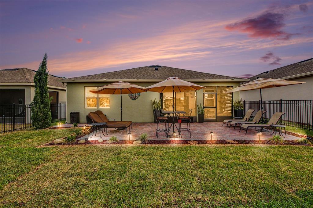 A backyard patio features brick pavers, lounge chairs, umbrellas, and a grassy lawn at sunset — 17651 BLAZING STAR CIRCLE, CLERMONT, FL