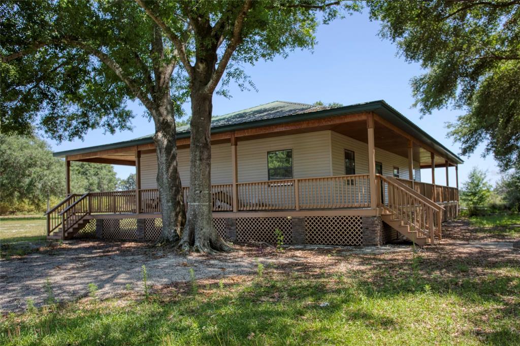 A tan house with a wrap-around porch and lattice skirting sits under large oak trees — 5525 MITCHELL BRIDGES ROAD, CLERMONT, FL
