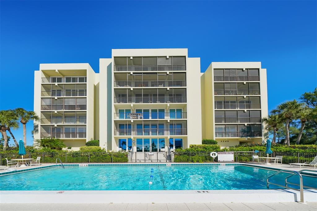 A multi-story apartment building stands behind a rectangular swimming pool under a clear blue sky — 3240 GULF OF MEXICO DRIVE UNIT B303, LONGBOAT KEY, FL