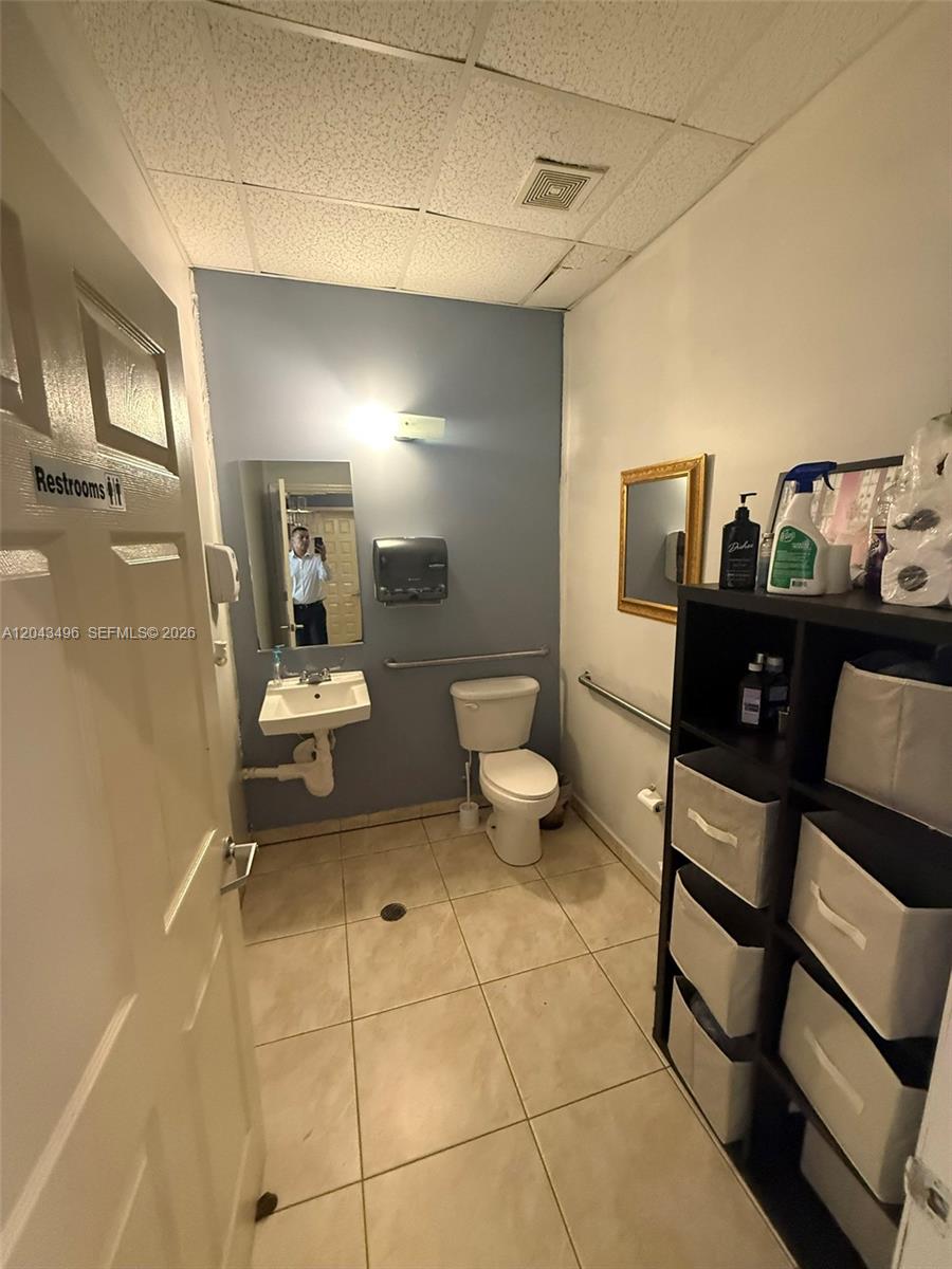 A small, tiled restroom features a toilet, a wall-mounted sink, and a storage shelving unit — 5921 NW 176th St # 9, Hialeah FL 33015, HIALEAH, FL